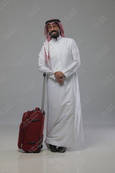A portrait of a smiling man wearing traditional Saudi attire, including a thobe and a shemagh. He is leaning on a red wheeled suitcase. The background is a neutral white.