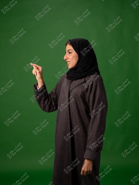 صورة لشخص يرتدي عباءة داكنة وحجابًا أسود, يشير بإصبعه نحو الجانب الأيسر. تم التقاط الصورة بخلفية خضراء واضحة.