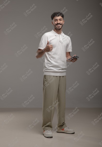 A portrait of a smiling man standing, wearing a white polo shirt and beige trousers. He is in front of a grey background, giving a thumbs-up with his right hand while holding a phone in his left hand. He is wearing white sneakers with orange accents.