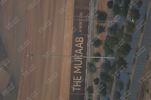 The image shows an aerial view of a construction site. There is a large sign that reads \"THE MUKAAB A NEW ICON\". The surrounding area appears desert-like with paved roads and rows of trees. A few cars can be seen parked on the road.
