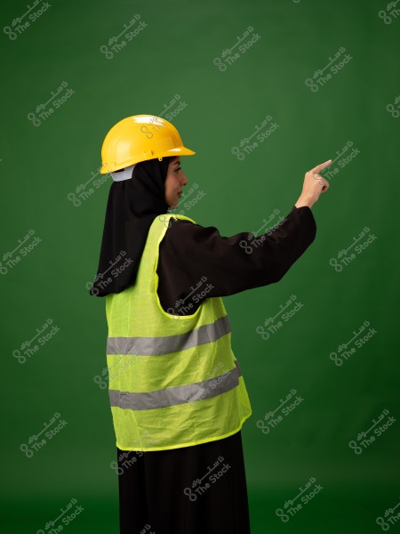 The image shows a woman wearing a yellow safety helmet and a green reflective vest, pointing with her finger in one direction. She is wearing a black abaya, and the background is completely green.