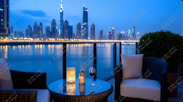 The image shows a nighttime view from a balcony overlooking a city filled with illuminated skyscrapers, possibly Dubai. In the foreground, there is a round glass table with a lit candle, a wine bottle, and a glass. A comfortable chair with white cushions is next to the table, and to the right, there is a green shrub.