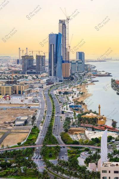 Aerial view of Jeddah city in Saudi Arabia showing the Red Sea Corniche road running along the coastline. On the left side of the image, there are modern high-rise buildings under construction and some completed ones with glass facades. On the right, a mosque with its minaret and traditional Arabic design is visible. The road runs straight, lined with palm trees, while the calm sea waters are seen to the right under the yellow sky.