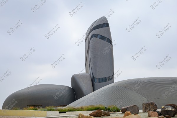 صورة لمبنى مركز الملك عبد العزيز الثقافي العالمي (إثراء) في الظهران, المملكة العربية السعودية. يظهر التصميم الفريد للمبنى بسطح معدني منحني يشبه الصخور, مع ناطحة سحاب مركزية ترتفع للأعلى. السماء ملبدة بالغيوم ويظهر بعض النباتات والصخور في المقدمة.