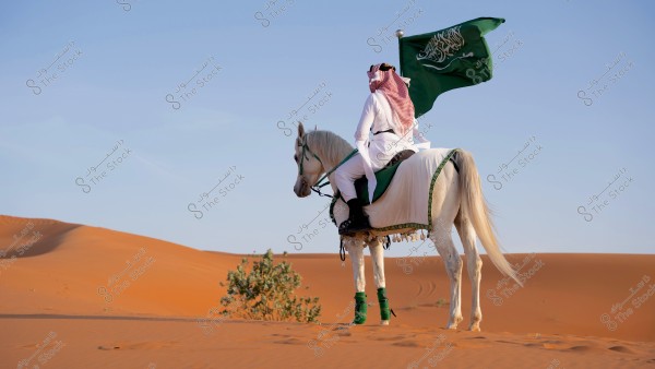 A man riding a white horse in the desert, holding a green flag. He is wearing traditional attire, including a white robe and a red-checkered headscarf. The scene suggests a desert setting in Saudi Arabia with sand dunes in the background.