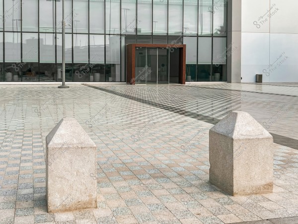 The image shows a wide outdoor plaza with light-colored, varied tiles on the ground. In the background, there is a modern building with glass walls and a large entrance door. A light post stands to the left, and there are square markers on the ground.