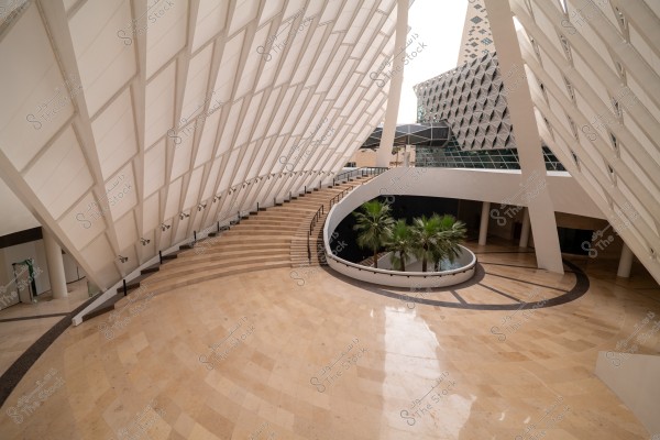 An image depicting a modern interior design of a building with innovative architecture, featuring curved white walls and a ceiling patterned with large squares. There is a semi-circular staircase leading to an upper level, surrounded by an open space with palm trees planted in the center. The design appears futuristic, with elements of glass and metal.