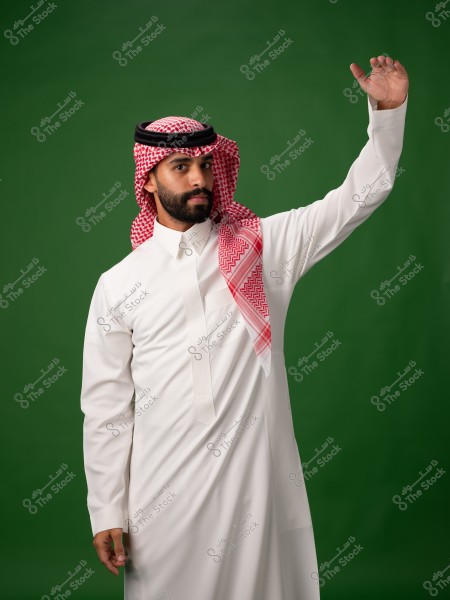 Image of a man wearing traditional Saudi attire, consisting of a white thobe and a red shemagh with a black agal. The man stands against a green background and raises one of his hands.