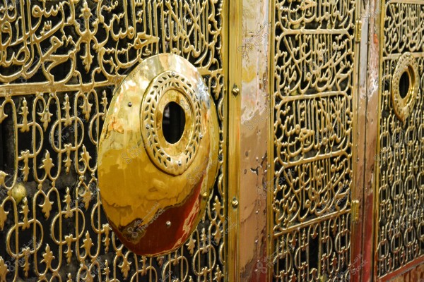 صورة لجزء من مشبك معدني مزخرف بتفاصيل دقيقة ونصوص عربية منقوشة بالخط العربي. المشبك مصنوع من معدن لامع باللونين الذهبي والفضي, ويحتوي على تفاصيل هندسية ونباتية.