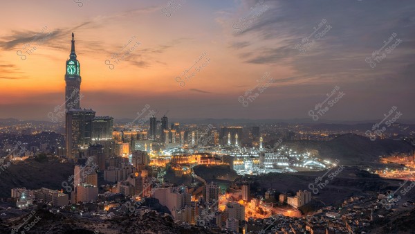 A panoramic view of Mecca at sunset, featuring the iconic clock tower on the left illuminated with bright lights. The center of the image is filled with the twinkling lights of buildings and homes. The horizon showcases warm hues of orange, pink, and pale blue, adding beauty to the holy city.