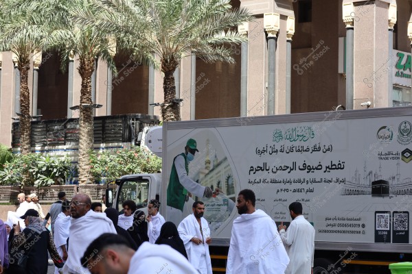 The image shows a group of people in white Ihram clothing standing in front of a truck with a board that says \"Iftar for the Guests of the Merciful in the Haram,\" alongside a diagram of the Haram. The truck has a picture of a person distributing food, with palm trees and surrounding buildings with Islamic architecture in the background.