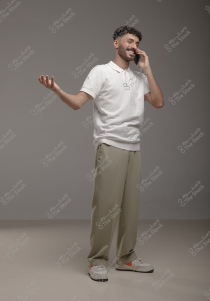 Image of a man standing in a studio wearing modern clothing. The man is wearing a short-sleeved white shirt, beige trousers, and white sneakers with orange accents. He is smiling and speaking on a mobile phone, gesturing with his other hand as if explaining something. The background is gray.