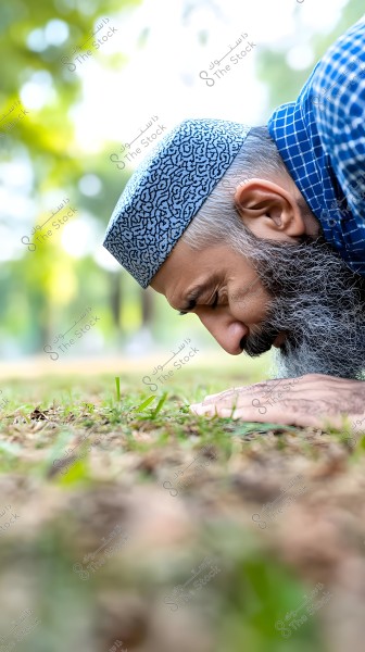 صورة لرجل يصلي في الهواء الطلق, يرتدي قبعة زرقاء مزخرفة وقميصًا أزرق بمربعات. الخلفية تحتوي على أشجار وضوء طبيعي مشرق.