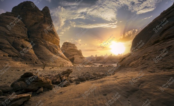 A natural landscape of a rocky desert with massive mountain formations under the light of the setting sun. The sky is adorned with glowing clouds, adding a dramatic atmosphere to the scene. Sparse desert vegetation is visible among the scattered rocks.