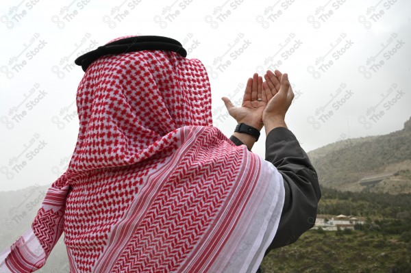 A shot of a Saudi man wearing the Saudi dress, praying and standing on one of the green mountains.