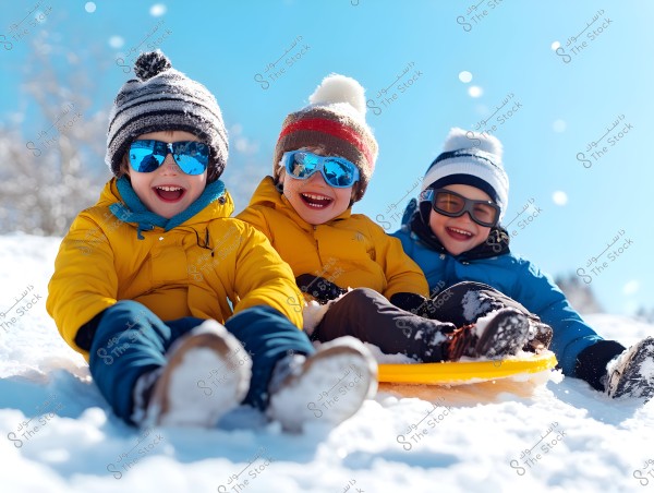 Three children sitting on a yellow sled in the snow under a bright blue sky. The children are wearing thick coats, two in yellow and one in blue, with winter hats and reflective sunglasses. They appear to be enjoying sledding and are laughing cheerfully.