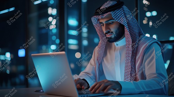 An image of a man wearing traditional Saudi attire, including a thobe, ghutra, and agal, sitting in front of a laptop in an indoor night setting with a background displaying city building lights. He appears focused and engrossed in his work on the computer.