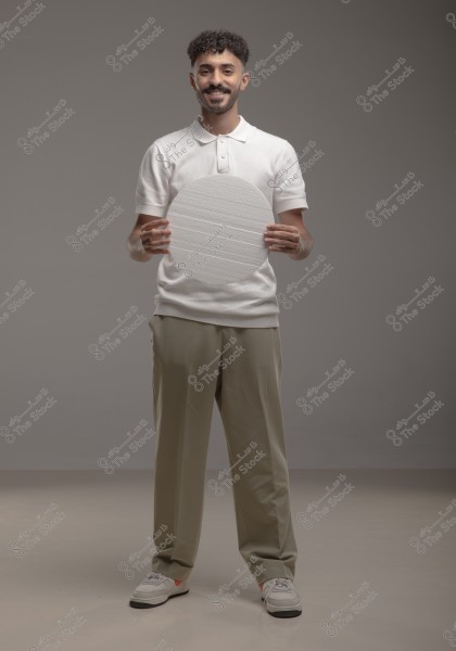 An image of a man standing against a gray background, wearing a white polo shirt and beige pants. He is holding a white circular board. The man has short, curly hair and is smiling pleasantly.