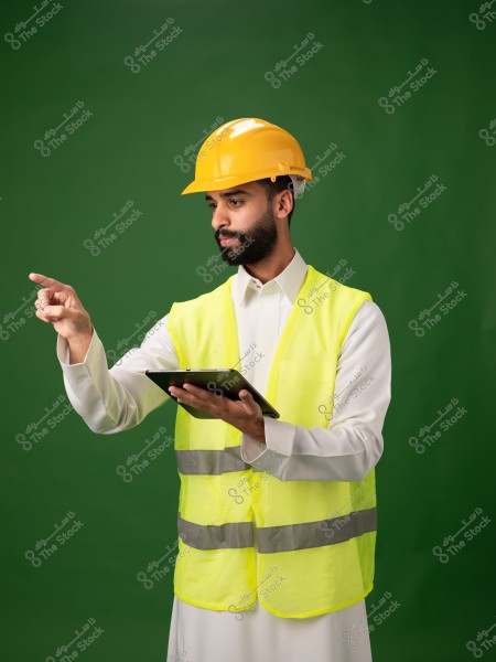 Image of a man wearing a yellow hard hat and a reflective safety vest over a traditional white garment, holding a tablet in one hand and pointing with the other. The background is green, highlighting his attire. His clothing suggests involvement in engineering or construction work.