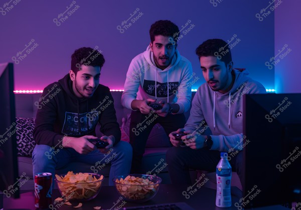 Three men sitting on a sofa in a room lit with neon blue and purple lights, enthusiastically playing video games, each holding a controller. In front of them is a table with bowls of potato chips and soft drinks. They are dressed casually in hoodies.