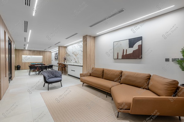 غرفة معيشة حديثة تحتوي على أريكة جلدية بنية اللون, وكرسي مريح, ولوحة فنية على الجدار. هناك منطقة طعام في الخلفية مع طاولة وكراسي, وجدار بواجهة رخامية.