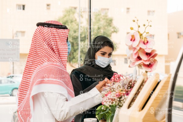 رجل سعودي يرتدي الزي السعودي التقليدي وكمامة و نظارة شمسية مع امرأة سعودية ترتدي العباءة والكمامة يقفان في المتجر عند الكاونتر للطلب