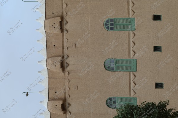 The image shows the facade of a traditional building in Najdi architectural style. The walls are made of mud, featuring arched green windows. Triangular designs can be seen along the upper edge of the wall, and part of a tree is visible at the bottom.