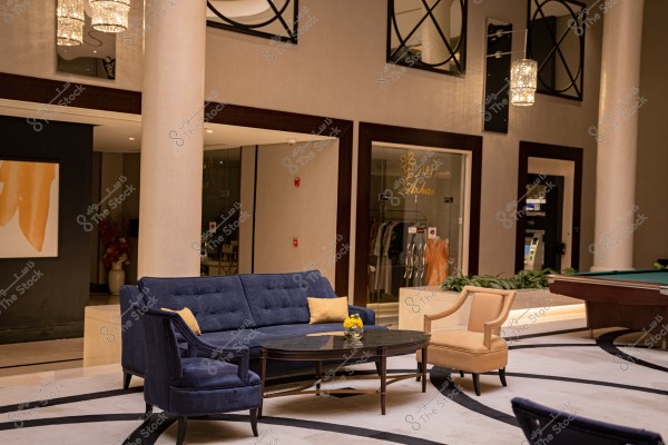 The image shows an elegant indoor seating area featuring a blue sofa with yellow cushions and armchairs in blue and beige around a circular coffee table. There is also a chandelier hanging from the ceiling and an art painting on the wall. In the background, a clothing store is visible through a glass window. The interior design reflects modern taste and luxury.