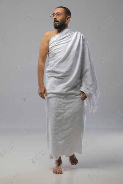 A photo of a man standing while wearing a white ihram, wrapping the white cloth around his body with the right shoulder exposed. He is wearing glasses and appears to be looking to the left. The image is taken against a simple gray background.