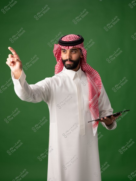 رجل يرتدي الزي السعودي التقليدي المكون من الثوب الأبيض والغترة الحمراء والعقال الأسود. يقف أمام خلفية خضراء ويمسك بجهاز لوحي بيد واحدة ويمد يده الأخرى للإشارة إلى الأمام.