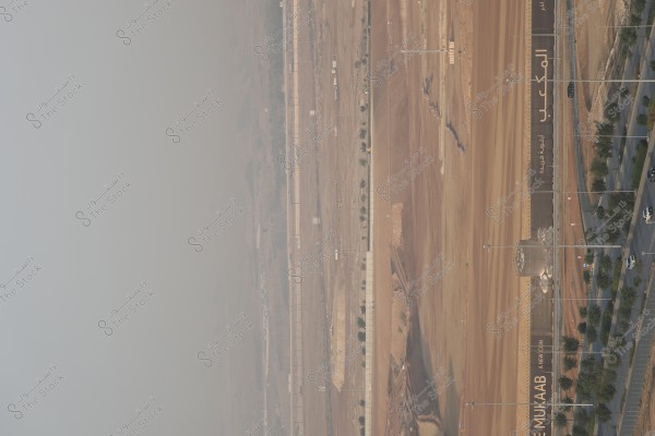تظهر الصورة منطقة صحراوية واسعة مع بعض الأعمال الإنشائية. يظهر في الجزء الأيمن طريق مزدحم بالأشجار على جانبي الرصيف مع عدة سيارات متحركة. هناك جدار أو حاجز عليه كلمة "المكعب" مكتوبة بالعربية والإنجليزية, مما يشير إلى وجود مشروع إنشائي مميز. الأفق يظهر بشكل ضبابي, مما يعطي منظرًا هادئًا للصورة.