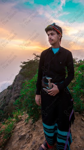 A Saudi man wearing a traditional dress at the top of the mountain and behind him clouds appear in Jizan