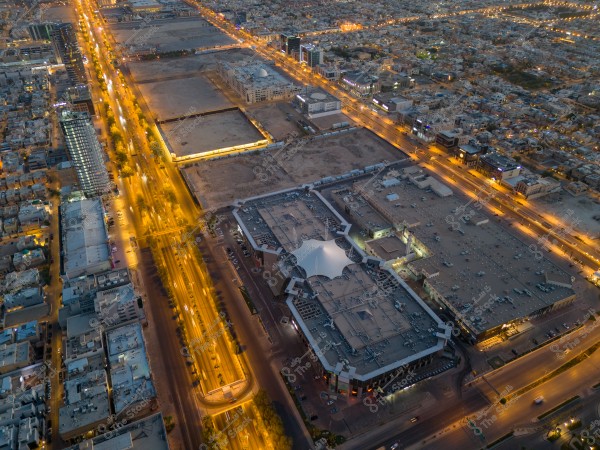 Aerial view of a city in the evening showing illuminated streets, buildings, and a large shopping area.