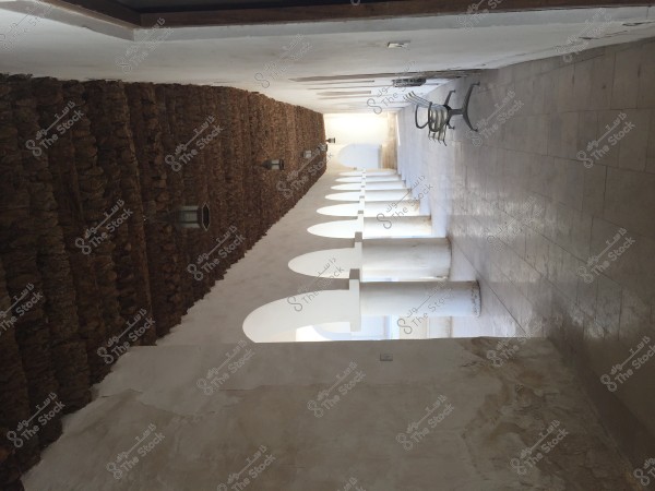 A long corridor in a traditional-style building, featuring stone walls and a clay ceiling, with white arched columns. Hanging lamps are attached to the walls, and on the right side, there is a row of metal chairs. Natural light streams through the arches, creating a serene atmosphere.