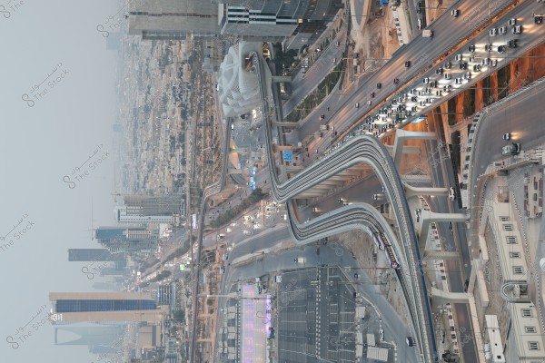 Aerial view of Riyadh city, showcasing an intricate network of intersecting highways in the city center. Cars can be seen traveling along the roads with tall buildings in the background. Modern architectural landmarks are visible, including a distinctive tower and glass buildings. The architectural style and urban scene suggest Riyadh, Saudi Arabia.