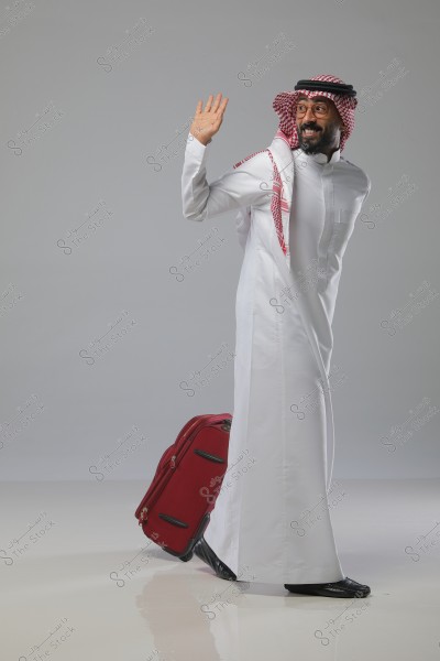 A man wearing traditional Saudi clothing, including a thobe, ghutra, and agal, waves his hand while pulling a red suitcase. He is standing in a studio with a gray background.