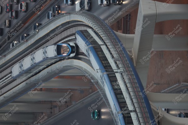 An aerial view of an urban scene featuring a modern light blue train traveling on an elevated bridge. Below the bridge, small and large cars are seen on a multi-lane road. The image highlights modern architecture in the design of the bridges and transportation infrastructure.