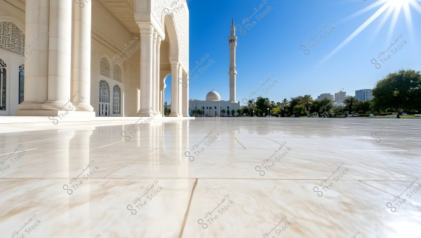 The image shows an open courtyard of a large mosque with white columns and Islamic architectural decorations. In the background, there is a tall minaret and a large dome. The sun shines brightly, reflecting on the glossy floor. Trees and plants are visible in the background along with a few tall buildings under a clear blue sky.