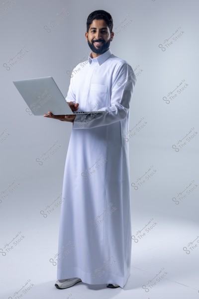 Image of a man wearing a white thobe, standing and holding a laptop. The man is smiling and has a beard, with a completely white background. The attire suggests the man may be of Gulf origin.