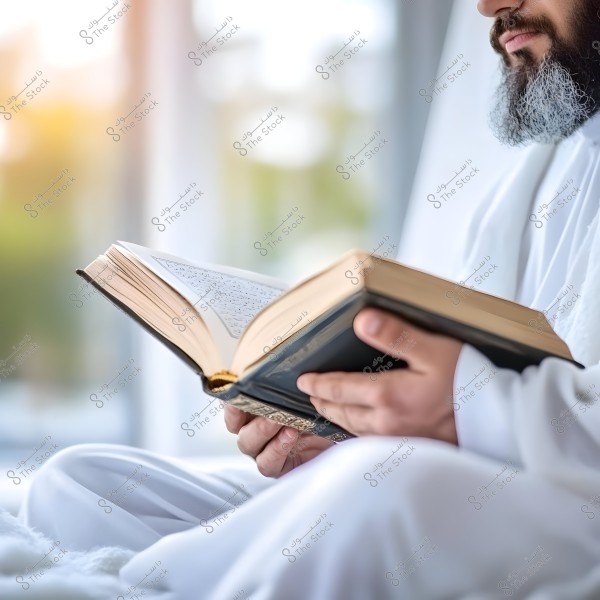 An image of a man wearing a white garment sitting and reading an open book, possibly the Quran. Part of his face and grey beard are visible. The background is blurred with natural lighting.