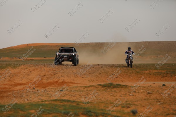 سيارة سباق ودراجة نارية تسيران على طريق رملي مفتوح في منطقة صحراوية, يظهر خلفهما غبار متطاير نتيجة الحركة السريعة. السماء ملبدة بالغيوم والأرضية تكتسي بطبقة خفيفة من العشب والأتربة.

-