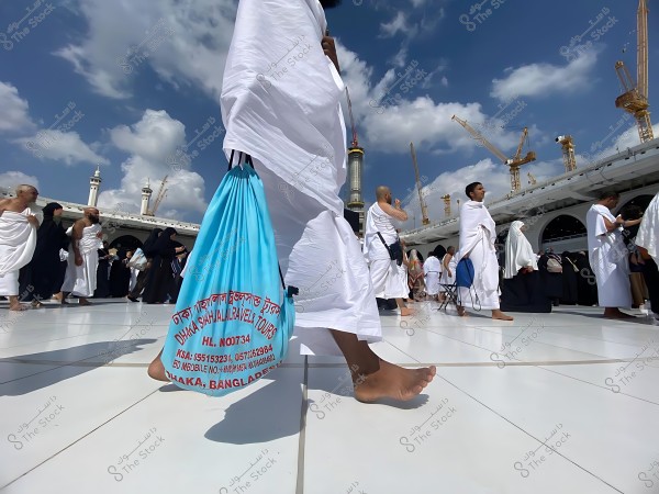 حجاج يرتدون ملابس الإحرام البيضاء يمشون في ساحة المسجد الحرام في مكة, تحت سماء زرقاء مع رافعات بناء في الخلفية.