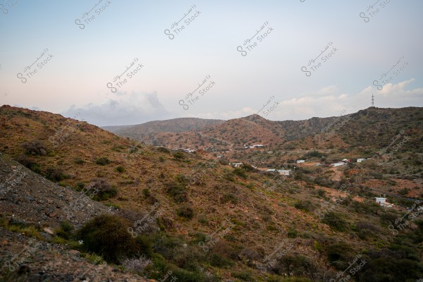 The image shows a natural landscape of a mountain covered with green grasses and small shrubs. Some small buildings are scattered across the mountain slopes, with a road winding through the scene. The sky is clear except for some clouds on the horizon, indicating a calm and  from Al Hada Taif natural atmosphere.