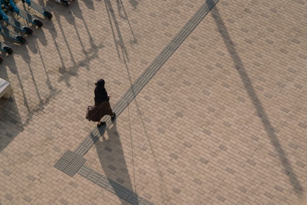 An overhead photo of a woman "with disabilities" in dark clothing walking on a brick sidewalk next to a row of electric scooters.