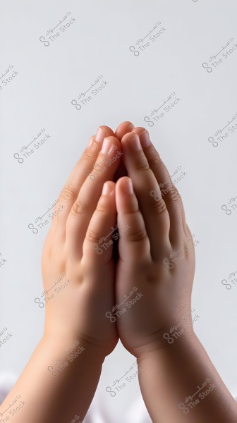 The image shows a pair of small hands held together in a gesture of prayer or supplication. The background is light gray, highlighting the smooth texture and fine details of the skin.