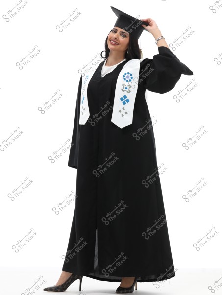 A portrait of a female graduate wearing a black graduation gown and a graduation cap, smiling. She has a white stole decorated with blue and gray patterns. She stands at an angle, holding the edge of the cap with her hand. The background is completely white.