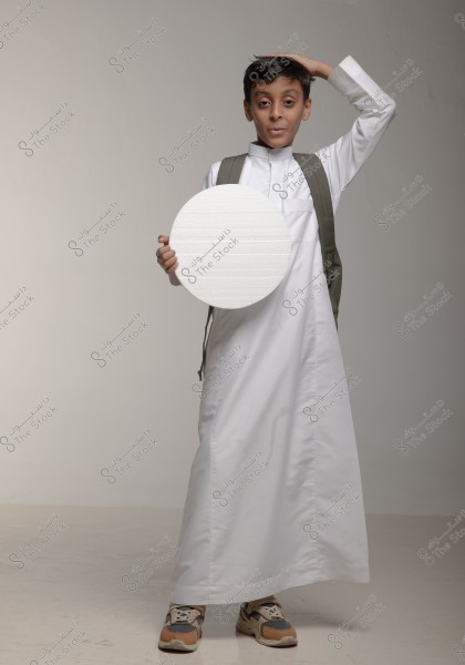 A young boy standing in a studio with a gray background, wearing a traditional white thobe and carrying a backpack. He holds a white circle in his left hand and places his right hand on his head. He is wearing comfortable athletic shoes. This portrait reflects a style of traditional clothing common in Saudi Arabia or Gulf countries.