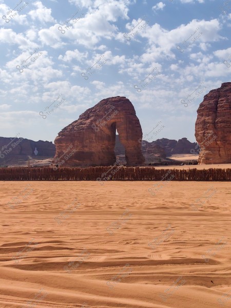 The image shows a large rock formation in a desert area with a blue sky dotted by light clouds. The rock is uniquely shaped, resembling an elephant, and is situated amidst red sands.