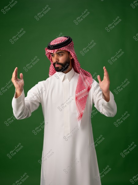 A portrait of a man standing in traditional Saudi attire, wearing a white thobe and a red ghutra with a black agal. His hands are raised. The background is green.