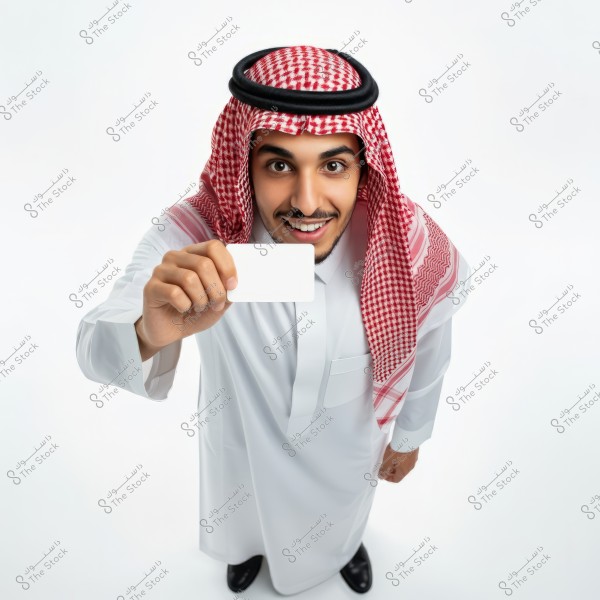 Image of a man wearing a white thobe and a red-checkered shumagh with a black agal. He appears to be holding a white card towards the viewer and is smiling. The clothing suggests he might be from Saudi Arabia or another Gulf country. The background is white.
